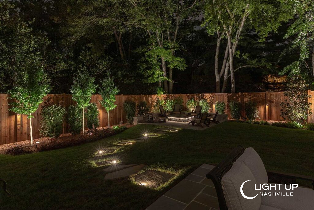 Profeslandscape lighting Nashvillesionally designed backyard landscape lighting featuring illuminated trees, stone walkway indicator lights, fence downlights, and strip lighting under a fire pit edge.
